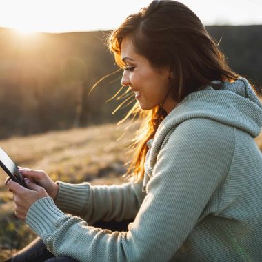 Smiling young woman using her smartphone while sitting on the ground in early spring sunny day and enjoying calm.